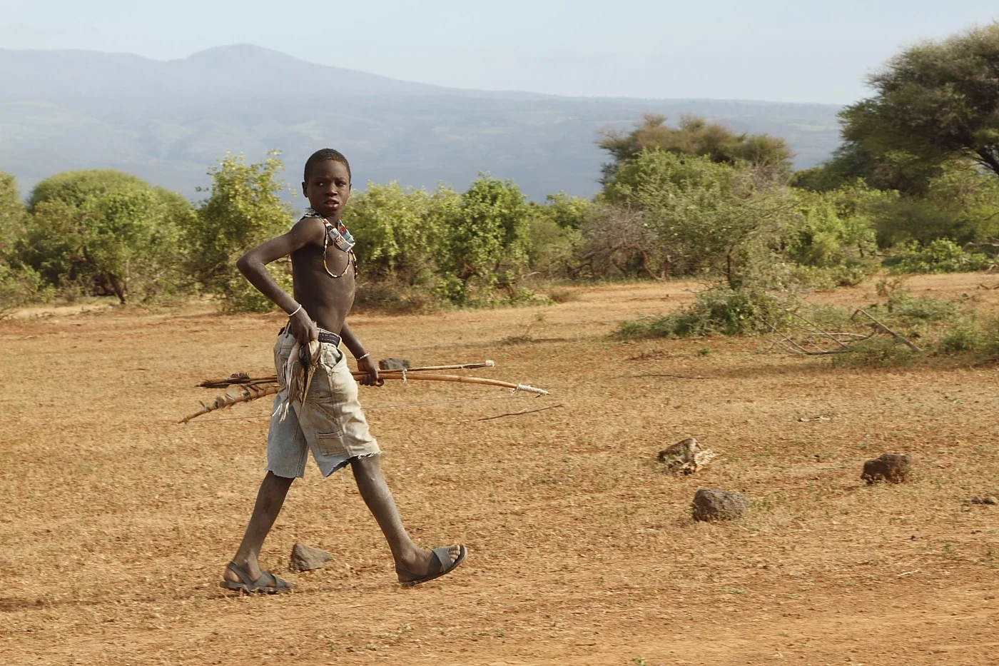 A young Hadzabe hunter walking across the dry ground with his bow and arrows