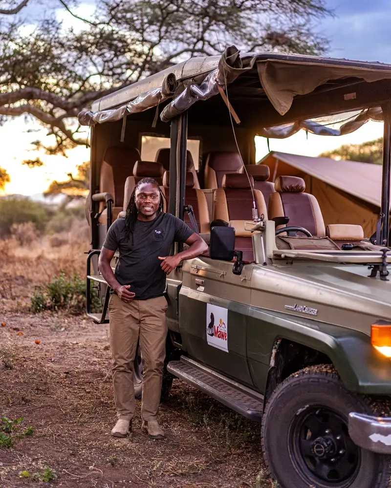 A safari guide standing next to an open-sided Toyota Land Cruiser safari vehicle parked near a tented camp ahead of a night game drive tarangire excursion.